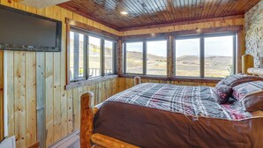 A wooden cabin bedroom with a large bed, a mounted TV, and a view of the landscape through large windows.