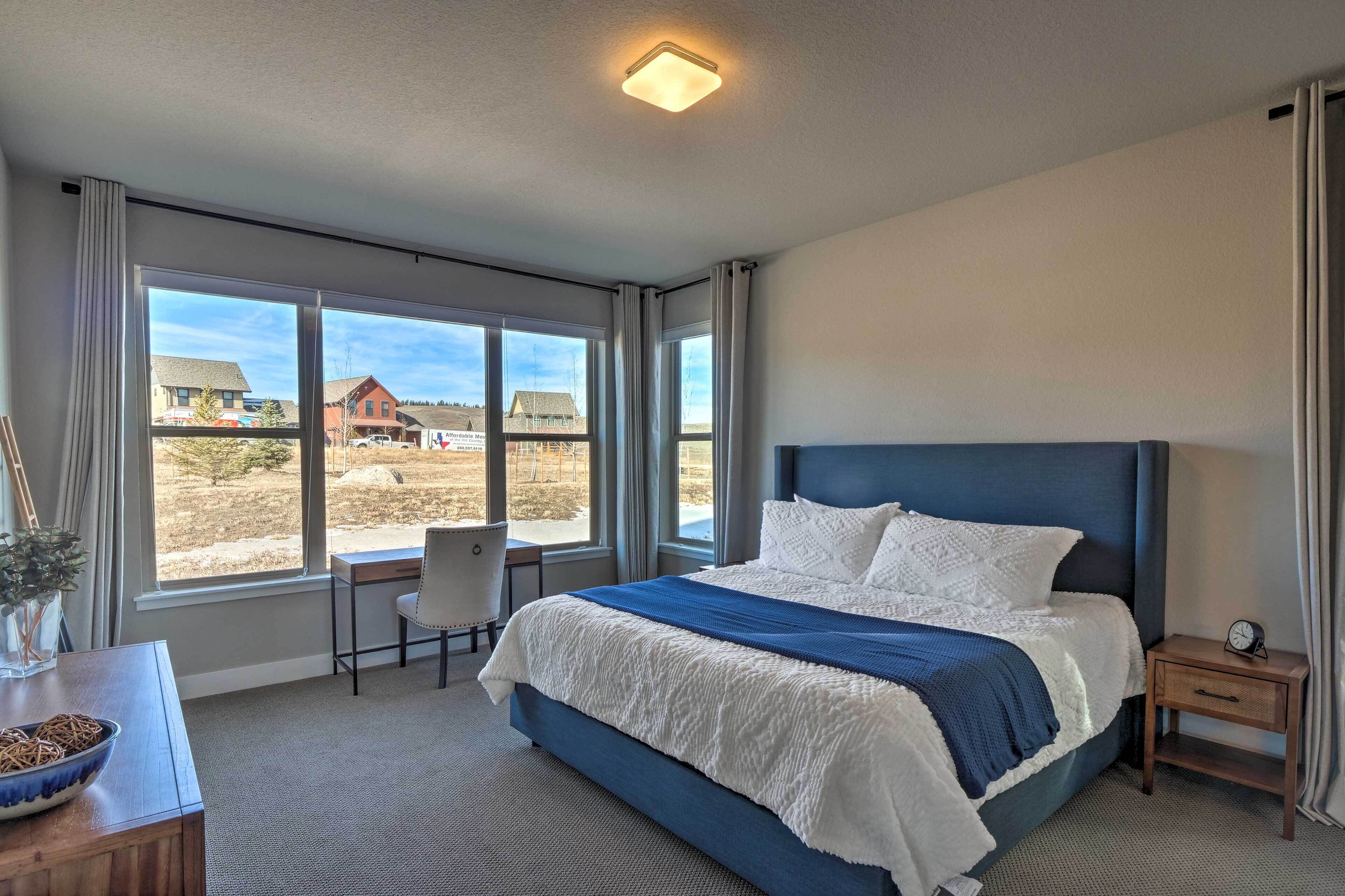 Un dormitorio con una cama grande, un escritorio, una silla y una ventana con vistas a casas.