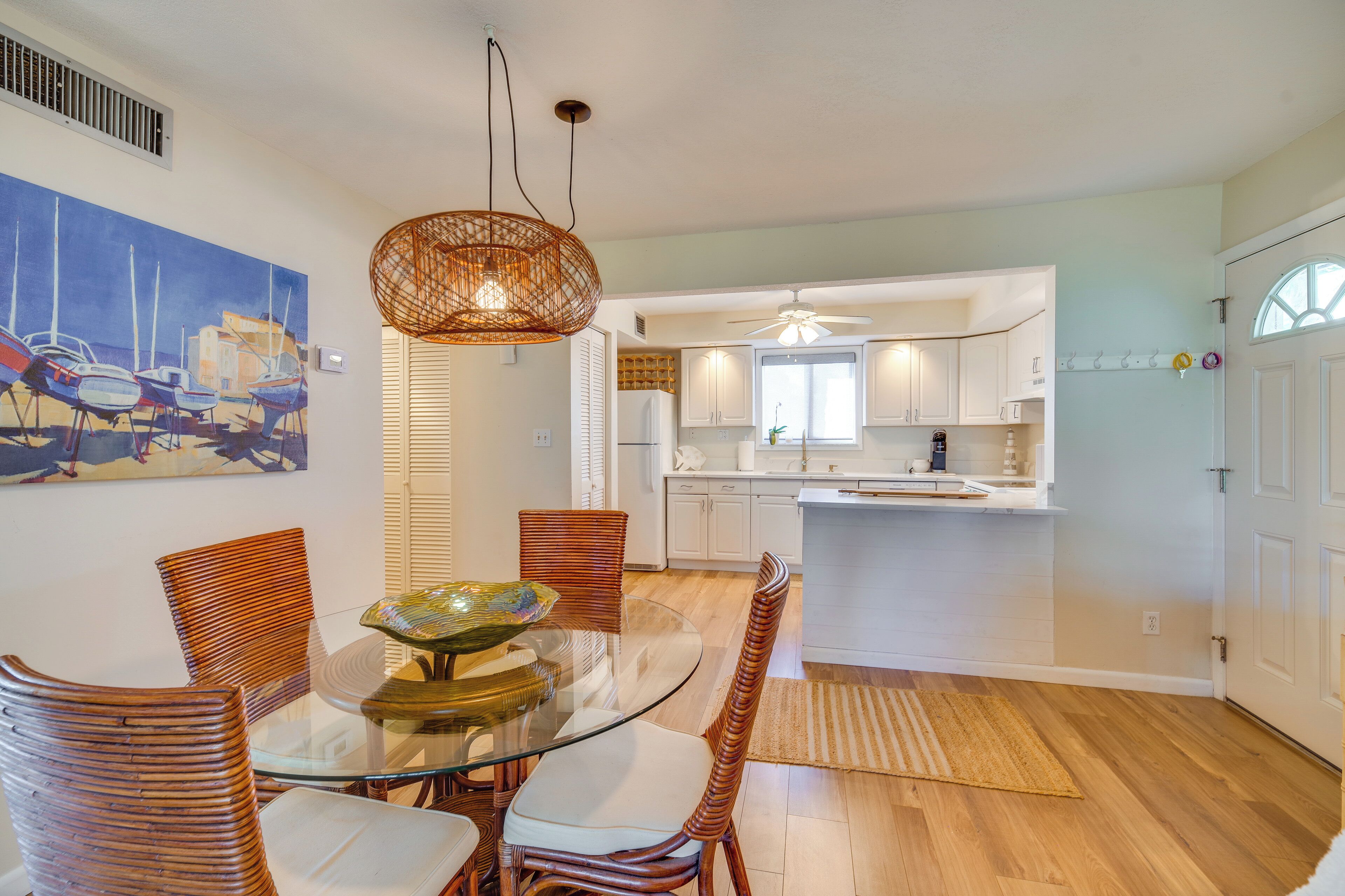 A dining area with a glass table and wicker chairs, a kitchen with white cabinets, and a painting of boats on the wall.