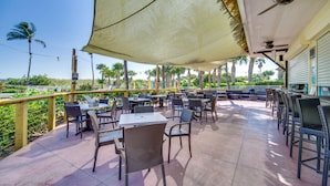 Une terrasse extérieure avec des tables et des chaises, un ventilateur de plafond et une vue sur des palmiers.