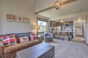A cozy living room with a brown leather sofa, plaid cushions, and a wooden coffee table. A kitchen with dark cabinets and a bar area is visible in the background. - Updated Studio Loft in Granby: Epic Mountain Views (Granby)