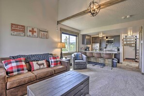 A cozy living room with a brown leather sofa, plaid cushions, and a wooden coffee table. A kitchen with dark cabinets and a bar area is visible in the background. - Updated Studio Loft in Granby: Epic Mountain Views (Granby)