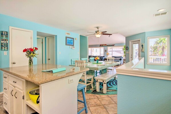 A kitchen with a granite countertop, white cabinets, and a vase with red flowers. There is a dining area with a wooden table and chairs, and a ceiling fan. - South Padre Home w/ Pool & Hot Tub: Walk to Beach! (South Padre Island)