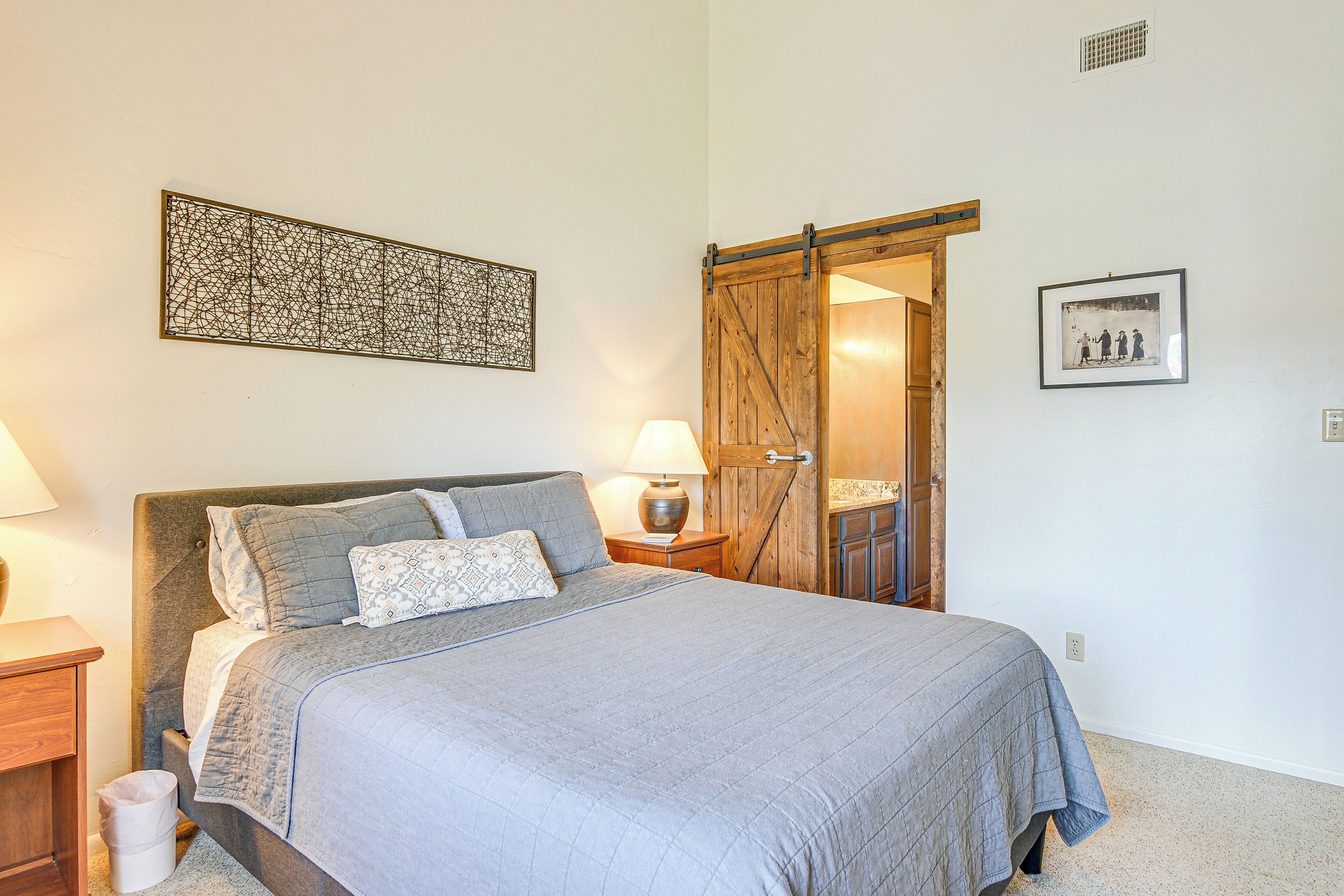 Een slaapkamer met een bed, nachtkastje, lamp, kunstwerk aan de muur en een deur naar een andere kamer.