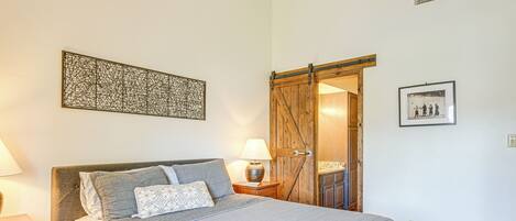 Een slaapkamer met een bed, nachtkastje, lamp, kunstwerk aan de muur en een deur naar een andere kamer.