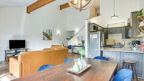 Amplia sala de estar con mesa de comedor de madera, sillas azules, sofá de cuero y una cocina con armarios oscuros y un refrigerador de acero inoxidable.