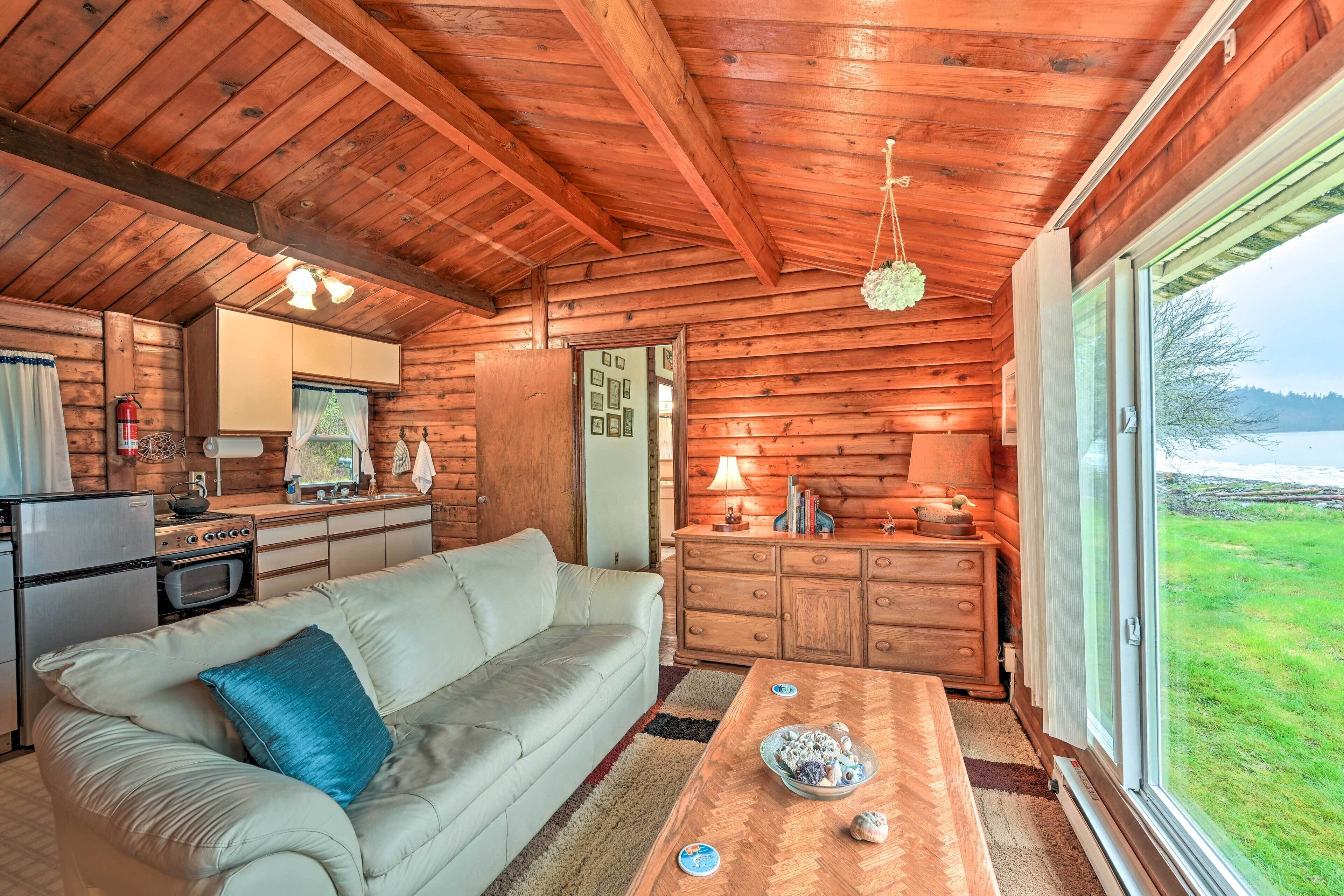 Ein gemütliches Blockhaus-Interieur mit Sofa, Esstisch, Küchenbereich und einem großen Fenster mit Blick ins Freie.