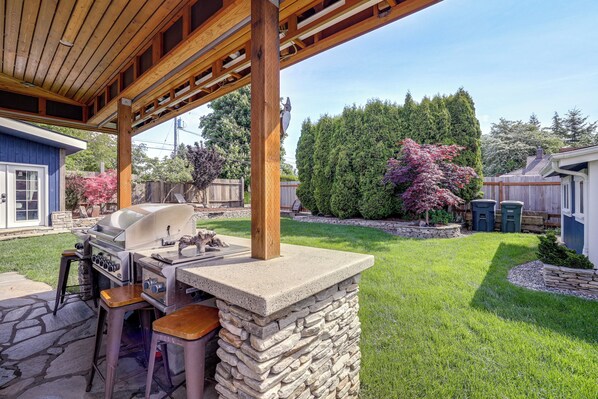 Un espace barbecue extérieur doté d’un grill en pierre et de tabourets, entouré d’une pelouse bien entretenue et surmonté d’une pergola en bois.