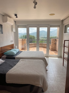 Quarto com duas camas, ventilador de teto e vista para uma varanda com parede de tijolos e vegetação.