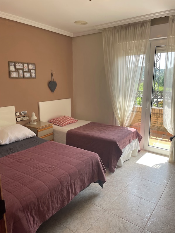 A hotel room with two beds, a nightstand, a desk, a chair, a window with curtains, and a view of greenery outside.