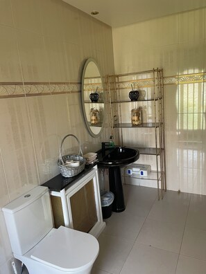 Casa de banho moderna com sanita, lavatório e espelho. Há prateleiras com objetos decorativos e uma cesta com toalhas.