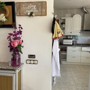 Uma cozinha com armários brancos, um porta-toalhas suspenso, um vaso com flores roxas e uma placa com a inscrição 'stay awhile'.