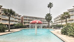 A swimming pool with a mushroom-shaped water feature, surrounded by palm trees and a multi-story building with balconies.