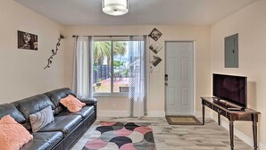 A living room with a black leather sofa, a TV on a wooden stand, a patterned rug, and a view through a window with curtains. - Ideally Located West Palm Beach Apartment! (West Palm Beach)