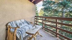A wooden bench and a sofa with a patterned blanket on a balcony with a wooden railing, overlooking a forested area.