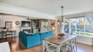 A spacious living room with a blue sectional sofa, a wooden dining table with white chairs, a TV mounted on a brick wall, and a large window with curtains. - 1 Mi to Main Street Sutter Creek: Poolside Oasis! (Sutter Creek)