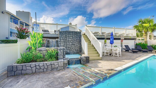 A swimming pool with a stone waterfall feature, a covered seating area with chairs and a table, and a white fence surrounding the property.