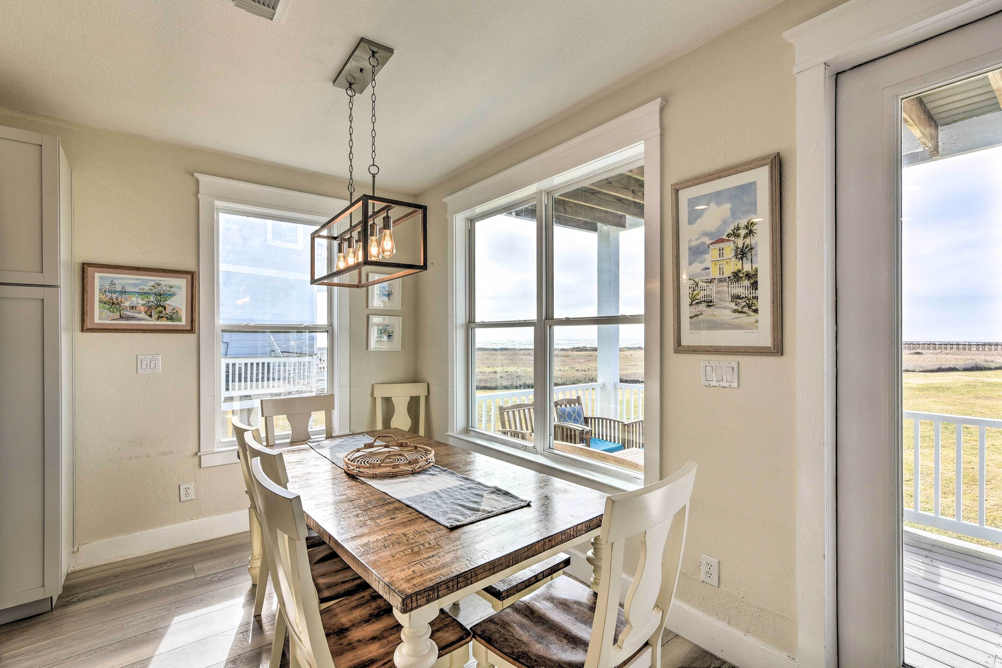 A dining area with a wooden table and chairs, large windows with a view of the beach, and a hanging light fixture.