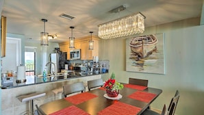 A modern kitchen with stainless steel appliances, a dining area with a large table and red placemats, and a chandelier above. - Idyllic PCB Condo w/ Pool Access & Ocean Views! (Panama City Beach)