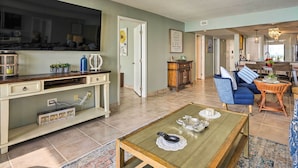 A spacious living area with a television, sofa, coffee table, and a kitchen in the background. - Idyllic PCB Condo w/ Pool Access & Ocean Views! (Panama City Beach)