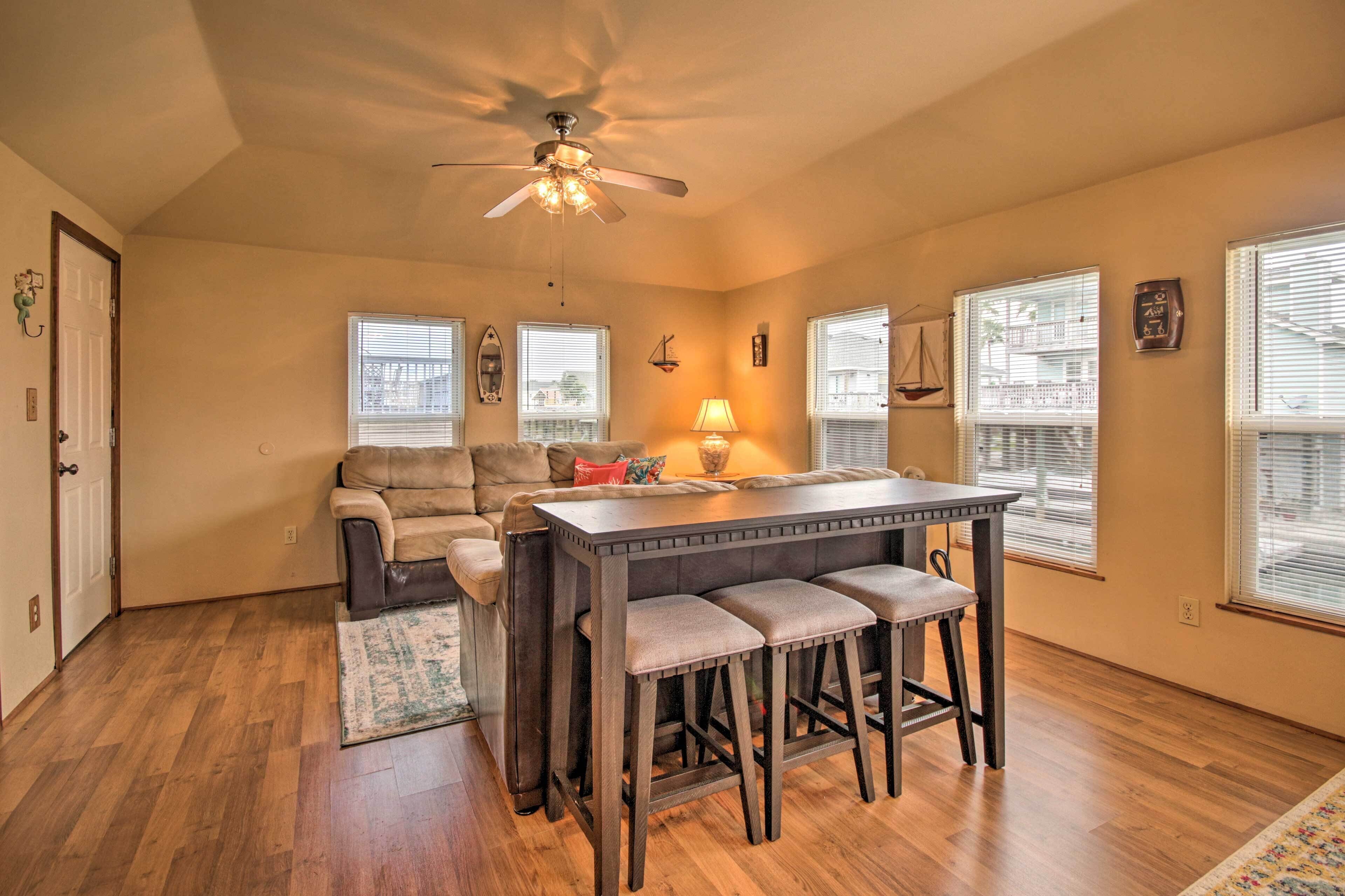 A spacious living area with a sofa, a coffee table, and a dining table with bar stools. There is a ceiling fan, a window with blinds, and a door.