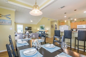 A dining area with a table set for four, a kitchen with wooden cabinets, and a living room with a television. - Walk to Galveston Beach: Coastal Escape w/ Yard (Galveston)