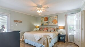 Een slaapkamer met een bed, een plafondventilator, kunst aan de muur, een nachtkastje en een raam met jaloezieën.