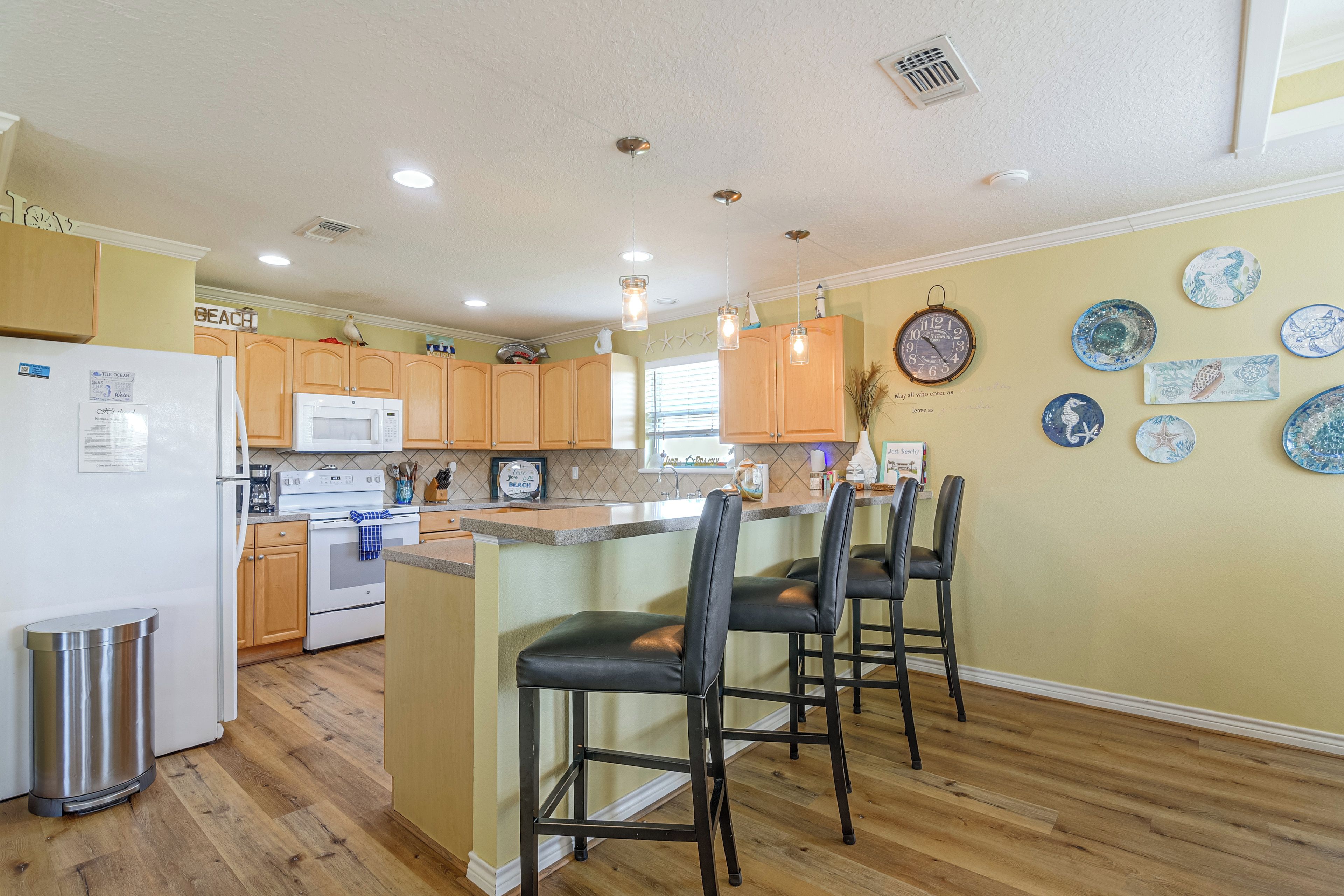 A kitchen with wooden cabinets, a white refrigerator, a microwave, and a stove. There are three black bar stools and a small trash can.