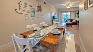 Uma sala de jantar com mesa de madeira posta para quatro pessoas, cadeiras brancas e decorações náuticas nas paredes.