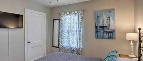 Une chambre à coucher avec un lit, un ventilateur de plafond, une fenêtre avec des rideaux à motifs, un tableau représentant des voiliers et une lampe de chevet.