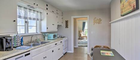 A modern kitchen with white cabinets, a dishwasher, a microwave, and a coffee maker. There is a window with blinds, a sink, and a rug on the floor.