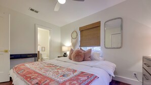 A bedroom with a large bed, a bedside table with a lamp, a dresser, a mirror, and a window with blinds. - 6 Mi to Dtwn Austin: Home w/ Screened Porch & Yard (Austin)