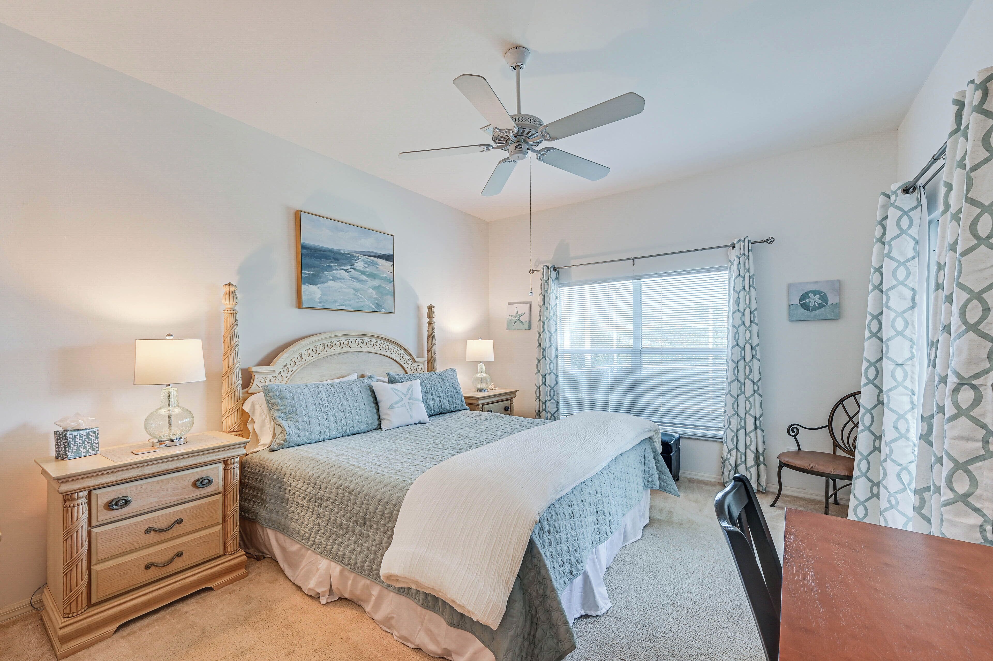 A bedroom with a bed, nightstands, a desk, a chair, a ceiling fan, and a window with curtains.