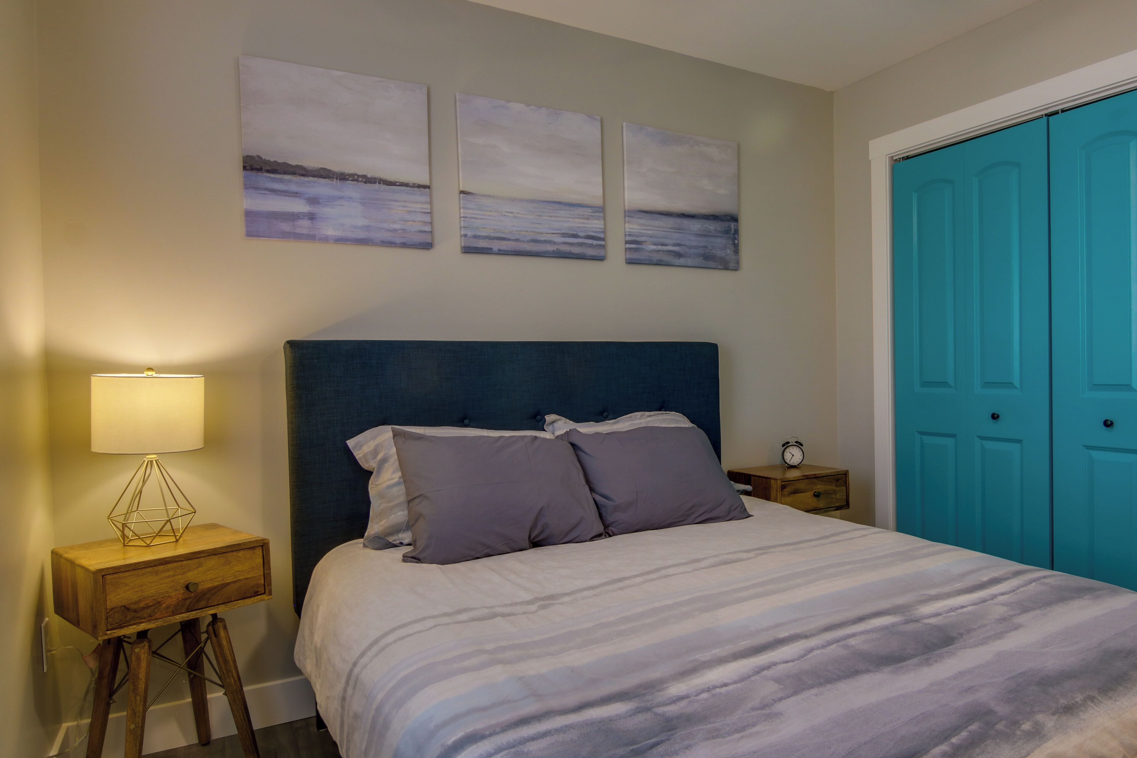 A bedroom with a bed, a nightstand with a lamp, a wooden drawer, and a blue door.