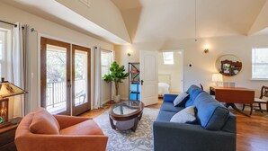 Ein geräumiges Wohnzimmer mit einem blauen Sofa, einem orangefarbenen Sessel und einem Glastisch. Es gibt einen Holzesstisch mit Stühlen, eine Lampe und einen Topfpflanzen.