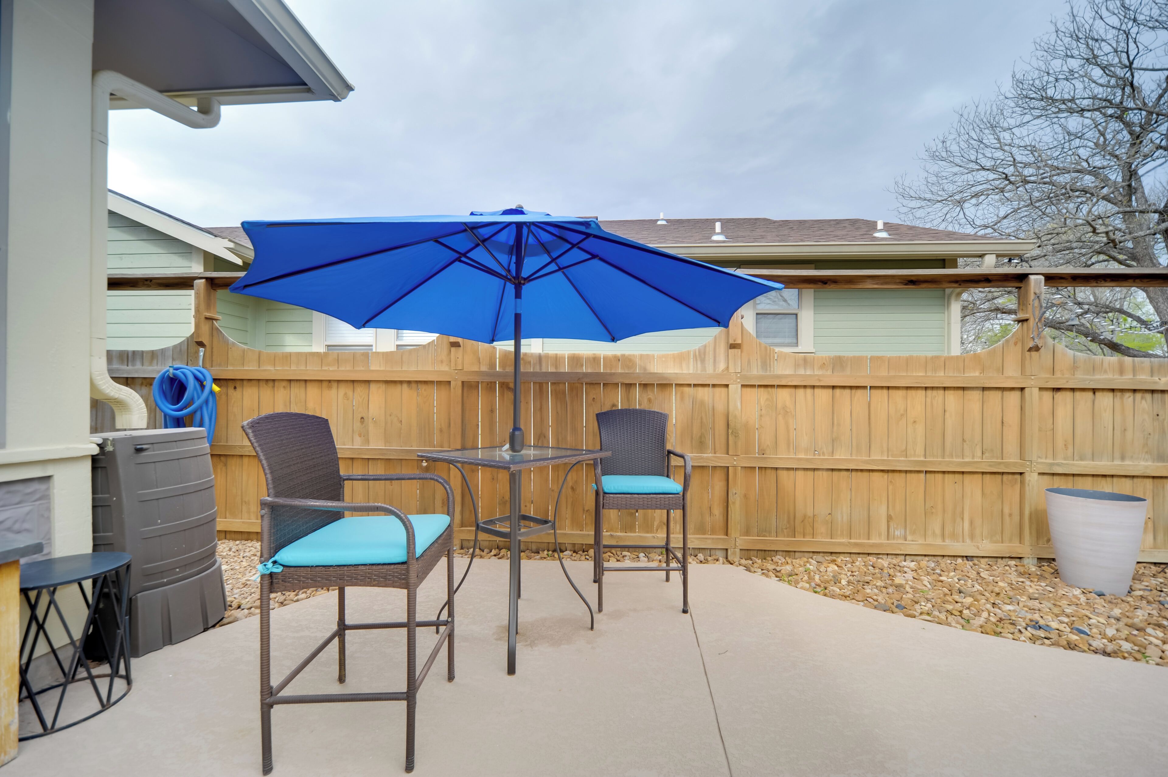 A patio with two wicker chairs and a table, a blue umbrella, and a wooden fence.