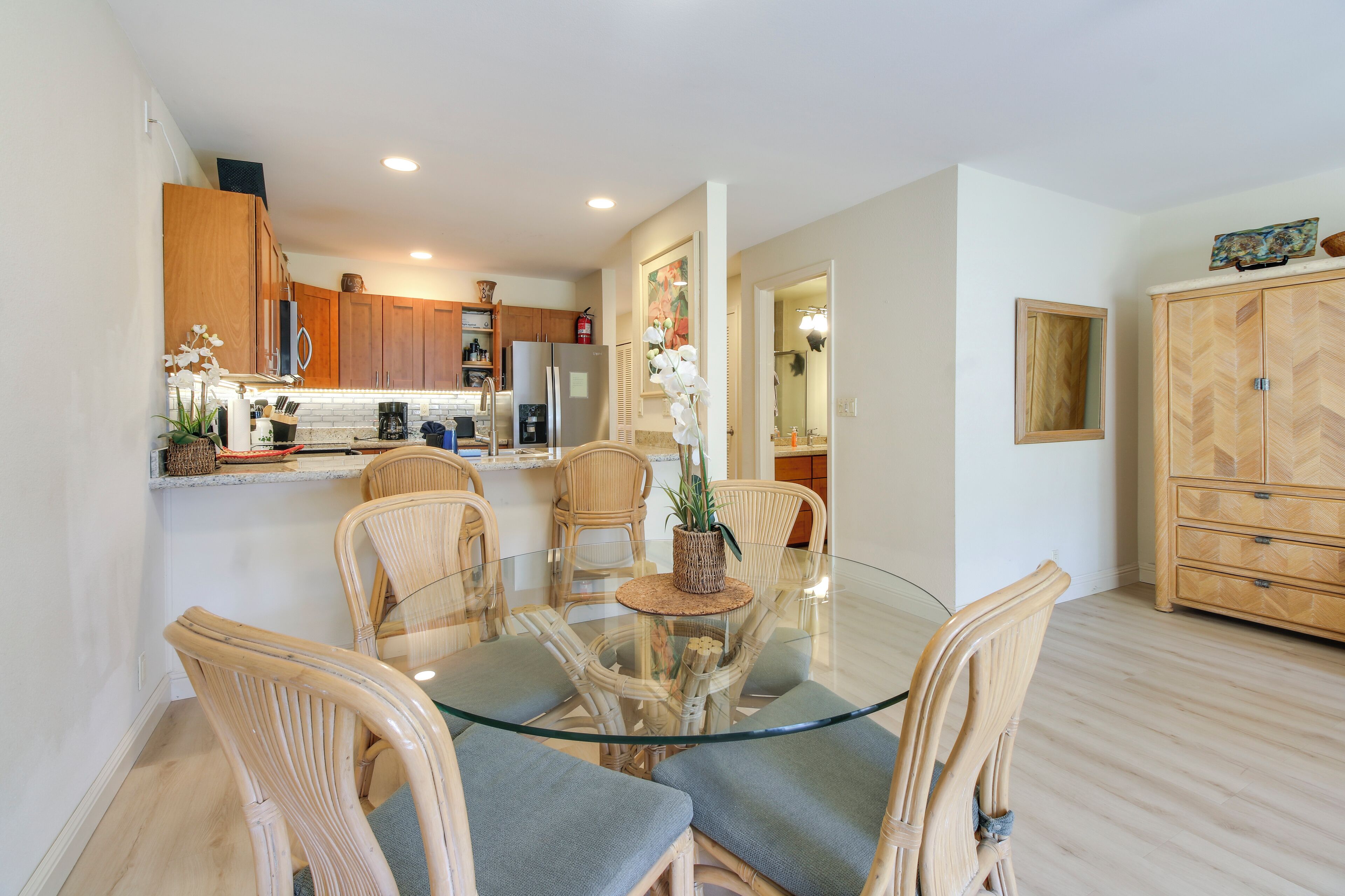 A modern kitchen with wooden cabinets, a glass dining table surrounded by wicker chairs, and a view into the dining area.