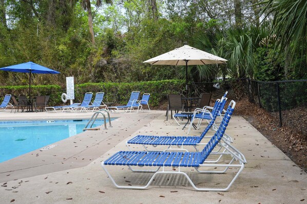 Un espace aménagé au bord de la piscine, avec des chaises longues et des parasols, entouré d’arbres.