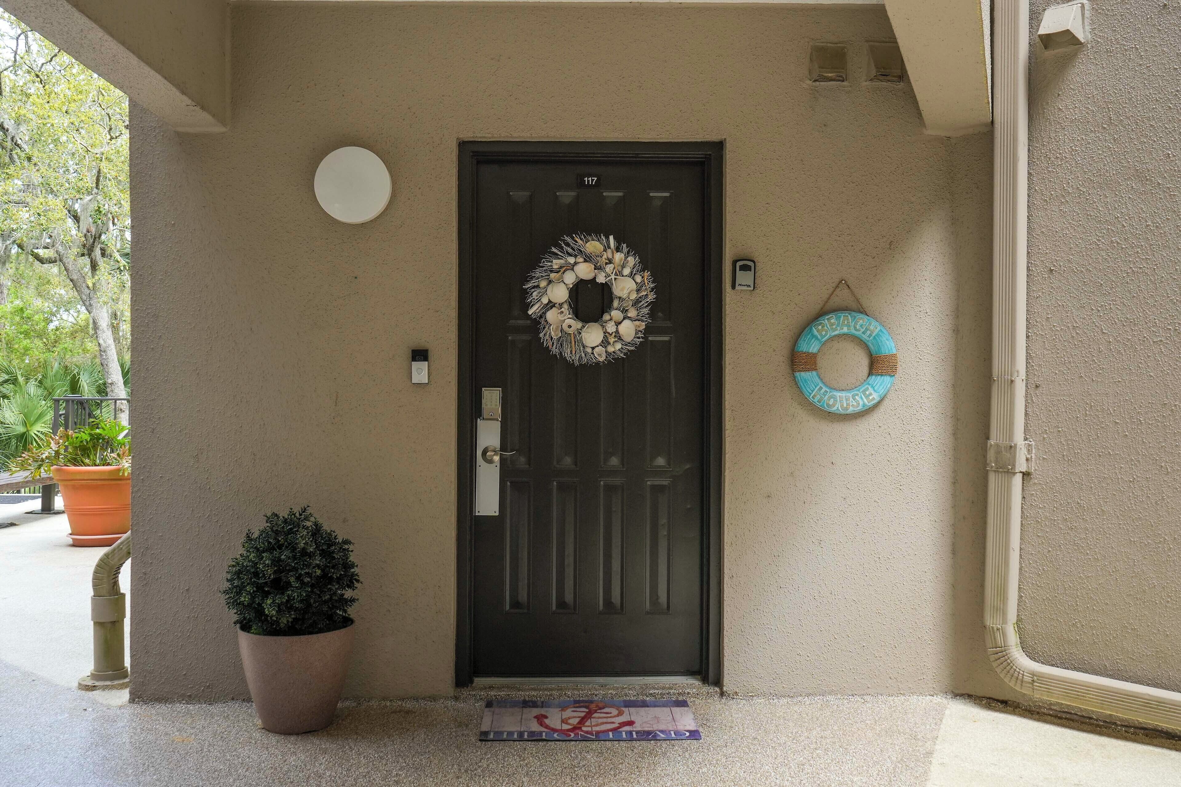 Une porte noire ornée d’une guirlande, un pot de fleurs et un cercle de sauvetage accroché au mur.