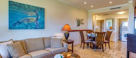 A living room with a sofa, coffee table, and a painting of a sea turtle. There is a dining area with chairs and a table.