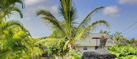 A pathway lined with palm trees leading to a house with a lion statue in front.