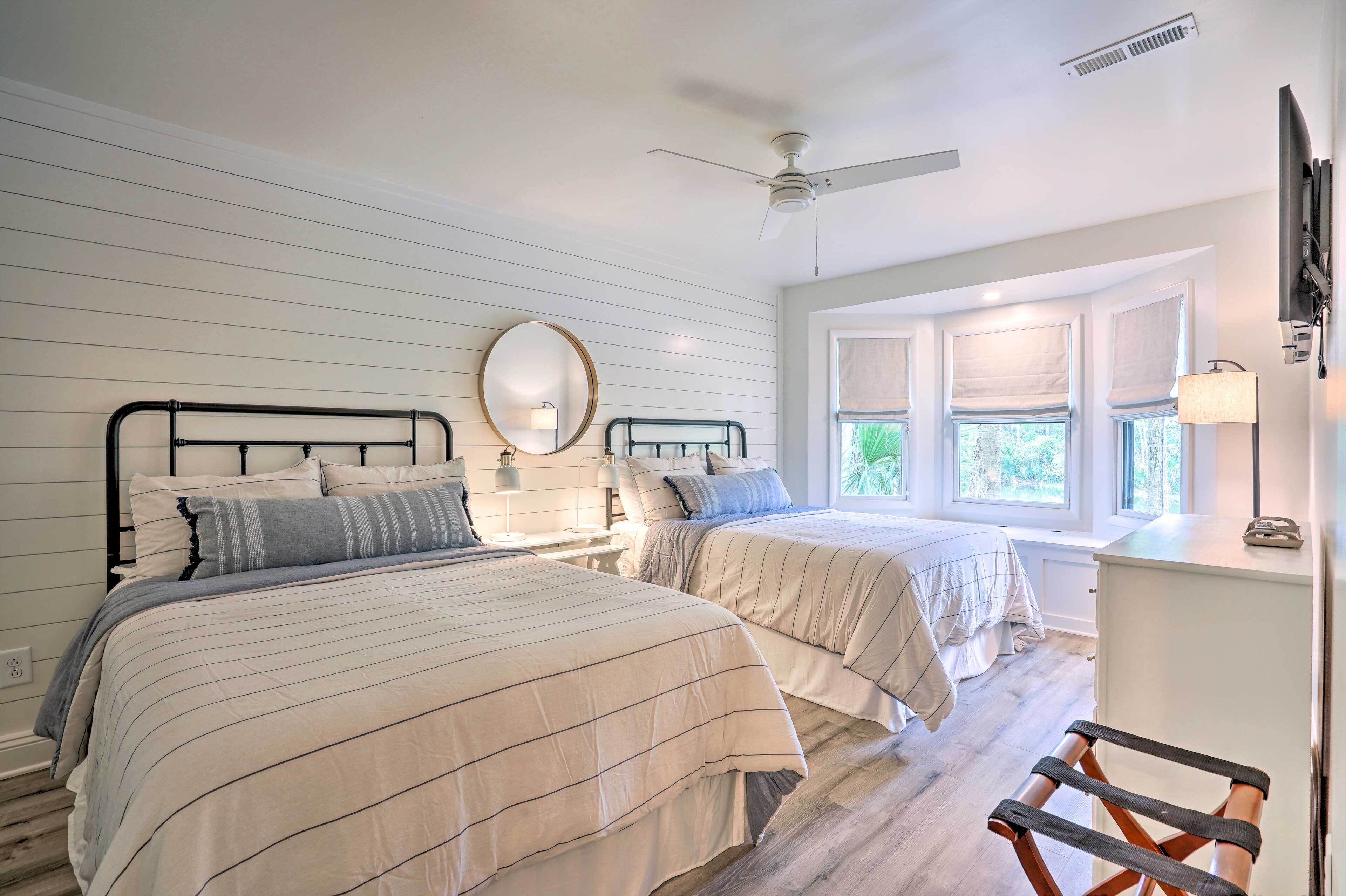 A bedroom with two beds, a ceiling fan, a window with blinds, and a desk with a lamp.