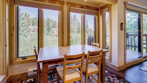 A wooden dining table with chairs, large windows, and a view of a house and trees. - Ski-in Breck Condo: Walk to Shops & Restaurants! (Breckenridge)