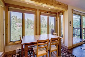 A wooden dining table with chairs, large windows, and a view of a house and trees.