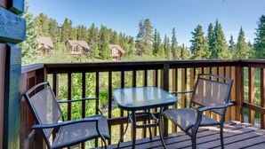 A wooden deck with a glass table and two chairs, overlooking a forested area with cabins. - Ski-in Breck Condo: Walk to Shops & Restaurants! (Breckenridge)