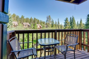 A wooden deck with a glass table and two chairs, overlooking a forested area with cabins. - Ski-in Breck Condo: Walk to Shops & Restaurants! (Breckenridge)