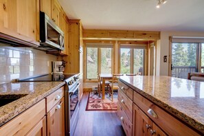 A kitchen with wooden cabinets, granite countertops, a microwave, and a dining area with a table and chairs. - Ski-in Breck Condo: Walk to Shops & Restaurants! (Breckenridge)