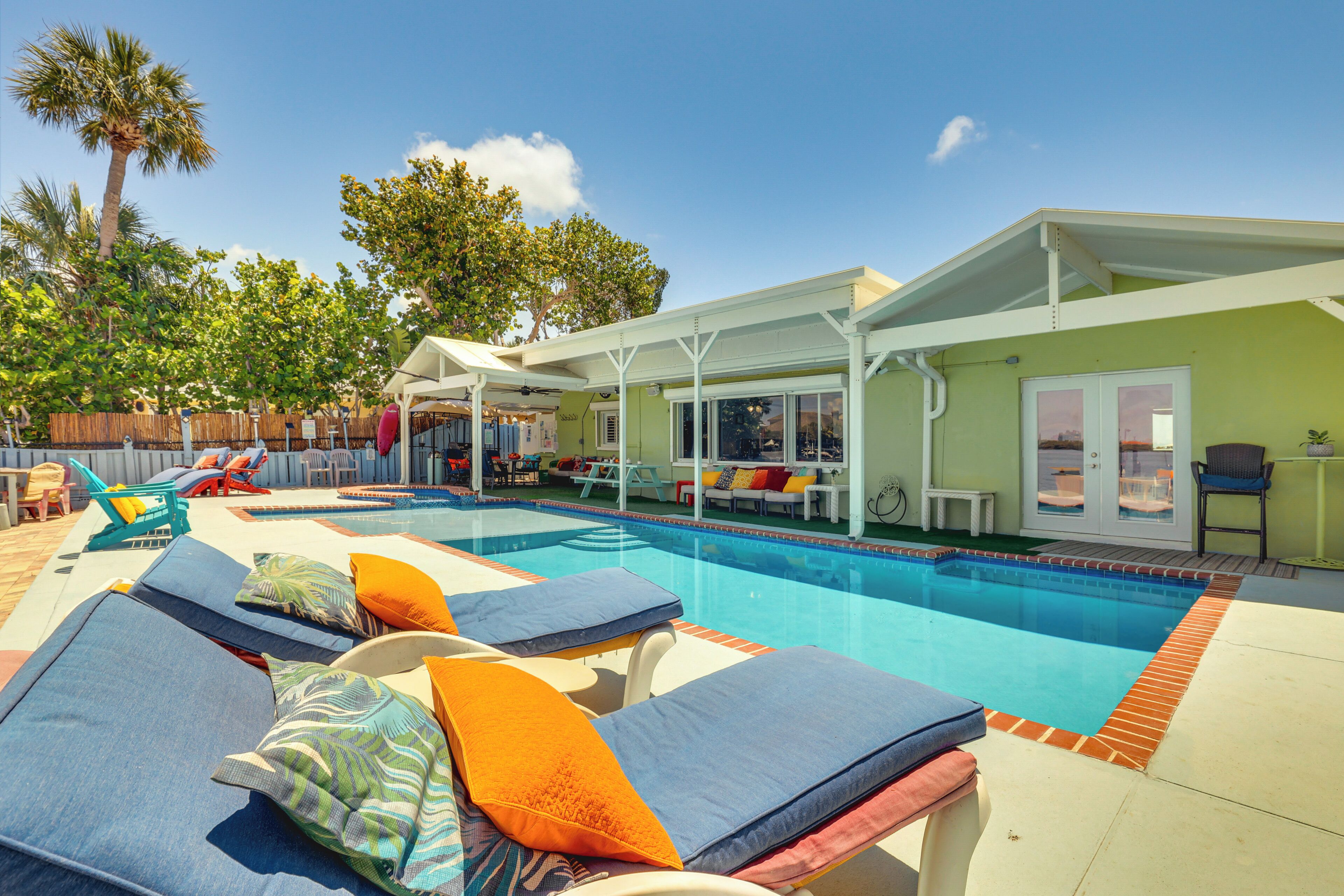 A swimming pool with lounge chairs and umbrellas, surrounded by a wooden fence and palm trees.