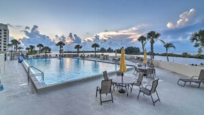 A poolside area with lounge chairs and umbrellas, a clear view of the ocean, and a sunset sky.
