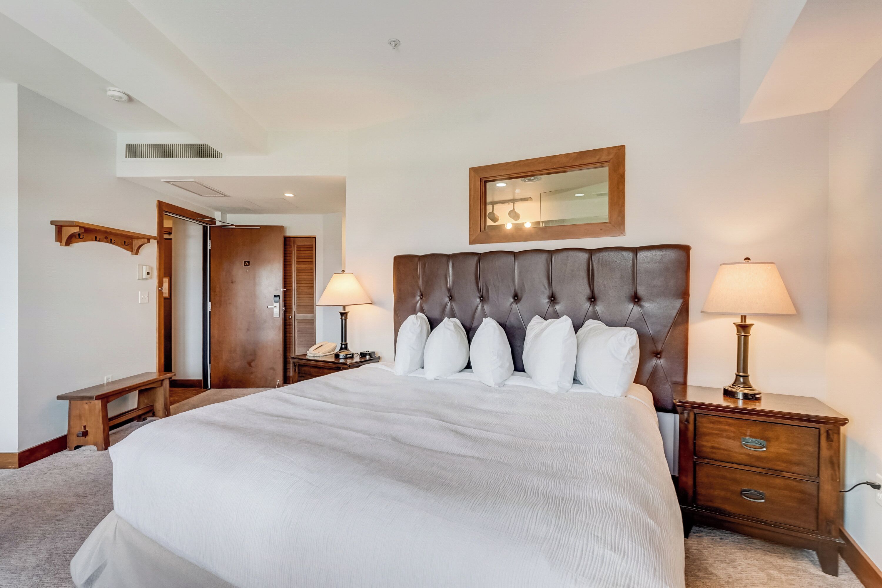 A neatly arranged hotel room with a large bed, two bedside tables with lamps, a wooden headboard, a mirror, and a bench.